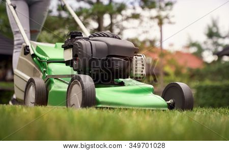 Close Up Of Female Legs Of Gardener Using Green Lawn Mower On Backyard. Selective Focus Of Woman Wor