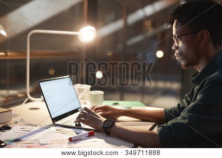 Serious African Man Sitting At The Table With Documents And Typing On Laptop Computer Till Late At O