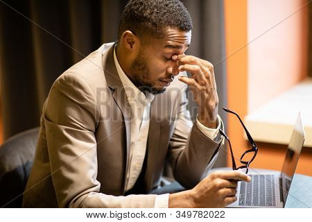Eyes Fatigue. Tired Afro Man In Suit Massaging His Nosebridge Holding Glasses Sitting At Desk With L