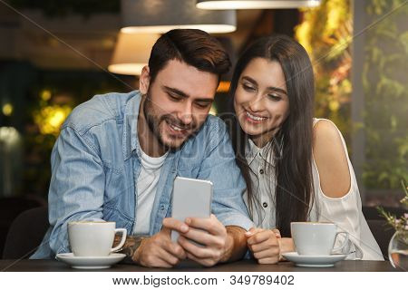 Young Couple Using Smatphone Sitting In Cafeteria Enjoying Coffee On Weekend Morning. Modern Dating 