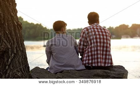 Male Teenager And His Father Sitting On Stone Near Lake And Talking About Life