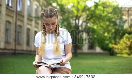 Beautiful Teenager Girl Reading Book On Bench In Campus, Preparing Homework