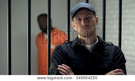 Prison Warden Looking To Camera Standing Near Cell With Imprisoned Afro-american