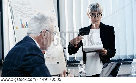 Businesswoman Pointing At Papers Near Colleague And Laptop In Office
