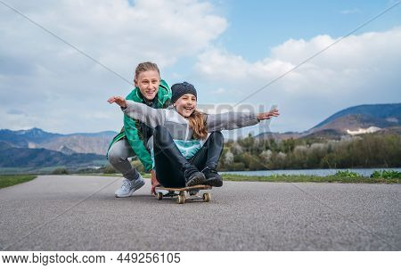 Laughing Kids Have Fun Using A Skateboard. Brother Pushing His Sister Sitting On A Skateboard And Ma