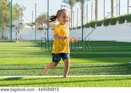 Cute Smiling Little Girl Running Across Green Sports Field.
