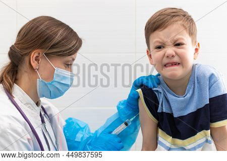 A Young Female Doctor In A Medical Gown And Mask Inoculates A Boy With A Syringe Against Coronavirus