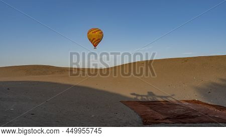 A Bright Red And Yellow Balloon Is Flying In The Blue Sky. A Carpet Is Spread Out On The Sand Dunes 