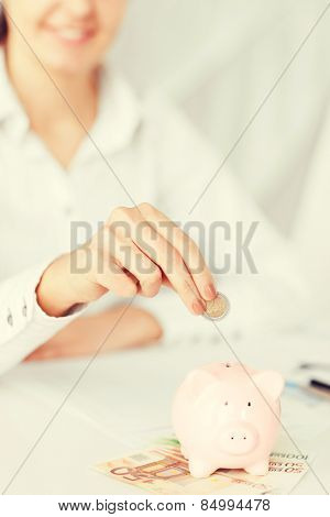 business, office, household, school, tax and education concept - woman hand putting coin into small piggy bank