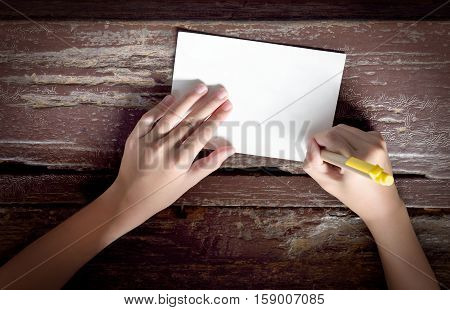 Top view of kid hand with yellow pen writing on paper note or postcard on old wooden table background. Flat lay of child's hands writing or drawing. Blank paper for copy space.