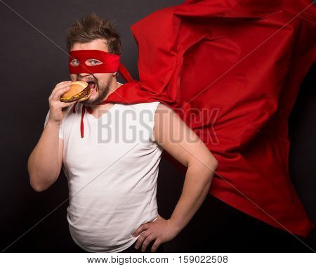 Super hero concept. Handsome man with red mask on eating hamburger while posing for photographer isolated on black background in studio.