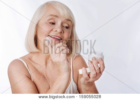 Fight with age. Joyful nice good looking woman holding a cream bottle and holding a finger while intending to put anti age cream on her face