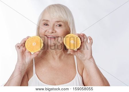 Two orange halves. Joyful charming senior woman holding two orange halves and smiling while standing against the white background
