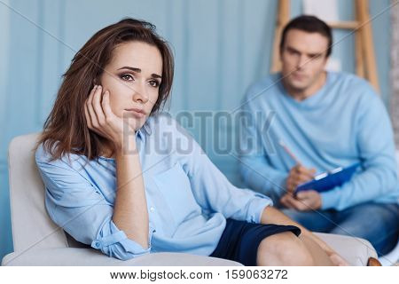 Troubled woman. Tires apathetic beautiufl woman holdign her hand on the chin and sitting in the chair while having an appointment with professional psychotherapist