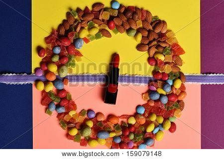 Red lipstick with purple ribbon and mix of dragee with raisins or peanuts inside marmalade or jelly candies almonds on colorful background children`s cosmetic