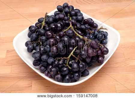 Still-life with lot of ripe black wine grape bunches with black berries in square bowl on wooden white background close up