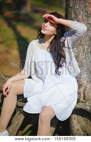 Young long-haired sweet girl in white dress and light sweater sitting near the tree and put hands to shield his eyes from the bright sun close up