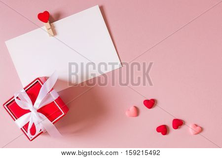 Valentine day composition: red gift box with bow stationery / photo template with clamp and small hearts on light pink background. Top view.
