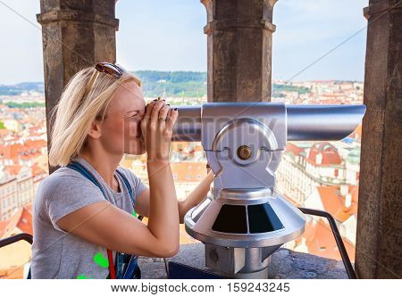 A young woman stands on top of the clock tower and looks at the Old Town in Prague using a telescope.