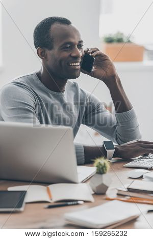 Always avaliable. Handsome young African man talking on the smart phone and smiling while sitting at his working place in modern office