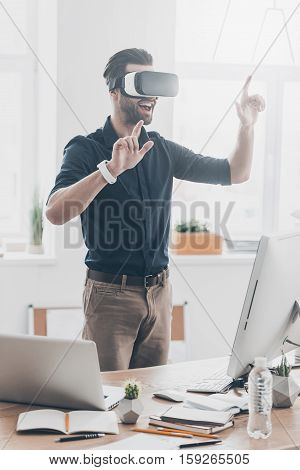 Everything is so real! Handsome young man in VR headset gesturing and smiling while standing in creative office