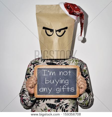 a young man dressing casual wear with a paper bag in his head and a santa hat in the top shows a chalkboard with the text I am not buying any gifts written on it