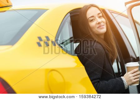 Cheerful woman with cup of coffee in hands sitting in yellow taxi with door open