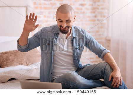 Hello there. Cheerful smiling bearded man sitting in the bedroom in front of the laptop while expressing happiness and waving hand