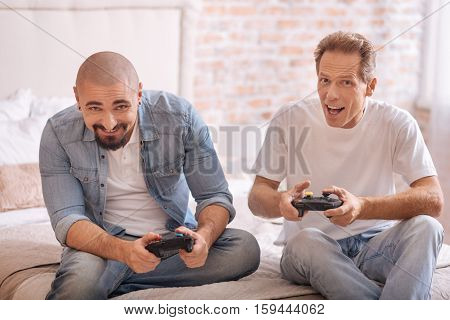 Full of fun. Delighted smiling male friends holding game consoles while sitting on the bed and expressing joy