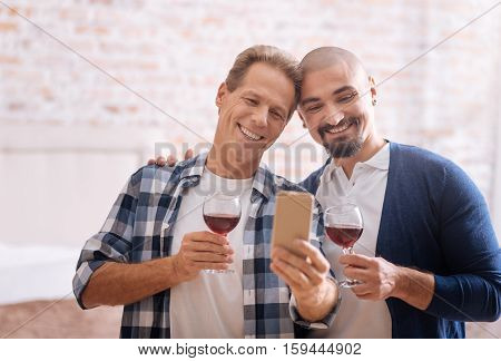 We enjoying our romantic moment together. Delighted cheerful gay couple standing in the bedroom and drinking wine while hugging each other and taking selfie