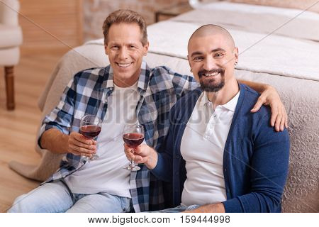 We love each other. Cheerful happy delighted non-traditional couple sitting in the bedroom and drinking wine while expressing love and care and hugging each other
