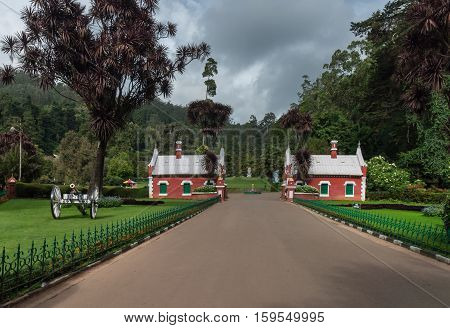 Nilgiri Hills India - October 25 2013: Wide gray walkway leads up to Heritage Gate at Ooty Botanical Garden. Small red houses against green background. Manicured lawns and cannon in foreground.