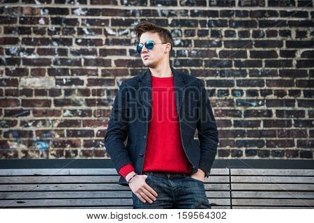 Fashionable young adult stylish man posing outdoors wearing red pulower jeans cotton jacket and sunglasses. Stylish handsome caucasian man with messy hairstyle.