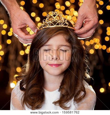 A little brunette girl getting crowned with a tiara in front of a starry background. She has closed eyes and looks pleased to be receiving her crown.