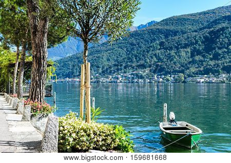 Vacation summer sunny lake view. Lonely wooden green boat on serene Lugano lake surrounded by hills green trees flowers blossoms and clear blue sky in Morcote Switzerland