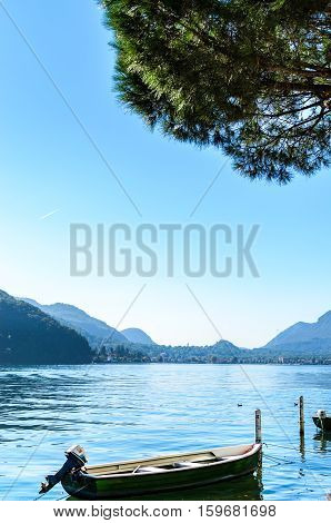 Vacation summer sunny lake view. Lonely wooden green boat under clear blue sky coniferous trees and beautiful background of serene Lugano lake surrounded by hills in Morcote Switzerland