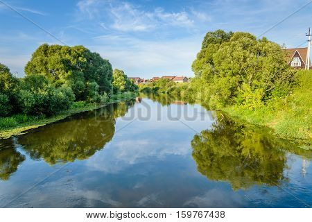 Summer warm evening. Riverfront Protva with footbridge. The village is in the background. Reflection of blue sky in the river water.