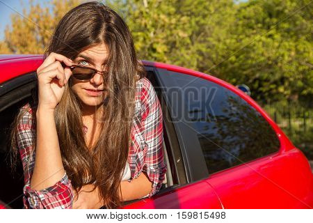 Portrait of happy young woman going on a road trip leaning out of window of the red car. She has blue eyes and brown hair. She has sunglasses on. She puts seductive eyes face showing.