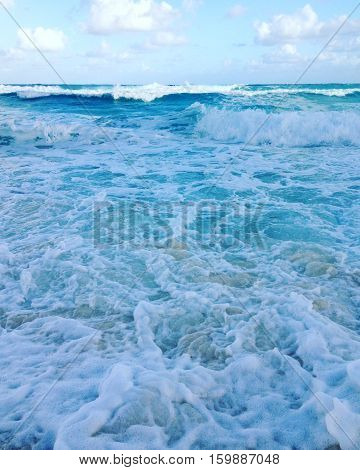 Blue and white ocean water waves with blue sky, clouds, sunshine and horizon background. Sea shore with sand, waves and sea foam.
