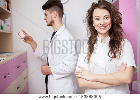 Smiling Doctor And Her Assistant Cheking Pills In The Background