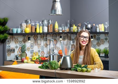 Portrait of young woman sitting on the table with fresh greens and blender at home