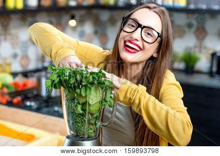 Young smiling woman pushing fresh greens into the blender in the kitchen at home. Healthy vegetarian smoothie for weight loss and detox