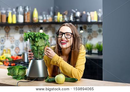 Portrait of young smiling woman with blender full of fresh greens, kiwi and gabbage ready to mix in the kitchen at home. Healthy vegetarian smoothie for weight loss and detox