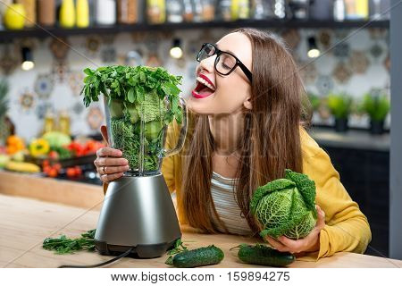 Portrait of young smiling woman with blender full of fresh greens, kiwi and gabbage ready to mix in the kitchen at home. Healthy vegetarian smoothie for weight loss and detox