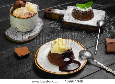 Chocolate lava cake (Molten) with ice cream on plate. Balls of ice cream in cup. Chocolate lava cake (Molten) with mint on board. Pieces of chocolate. Dark black wooden background.