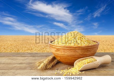 Bulgur or couscous in bowl. Bulgur in ceramic bowl and ears of wheat on table with field of wheat on the background. Golden wheat field and blue sky with beautiful clouds