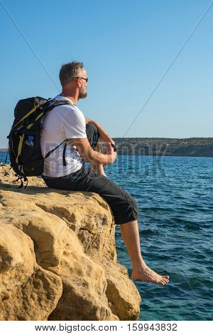Bearded Man With Backpack Sits On Rocky Shore Barefoot