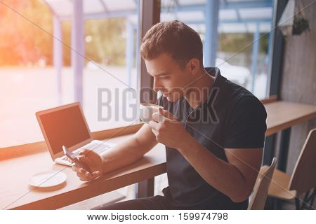 Man having a coffee break in modern cafe, while drinking a hot drink and browsing smart phone with laptop on table. Sun light effect