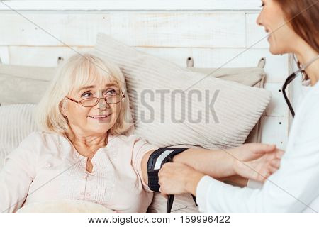Check it out. Pleasant elderly positive woma lying in bed while professioanl doctor measuring her blood pressure while visiting her patient at home