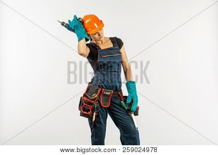 Sexy Workwoman In Overalls And Safety Helmet With Tool Belt Holding Electric Drill, On White Backgro